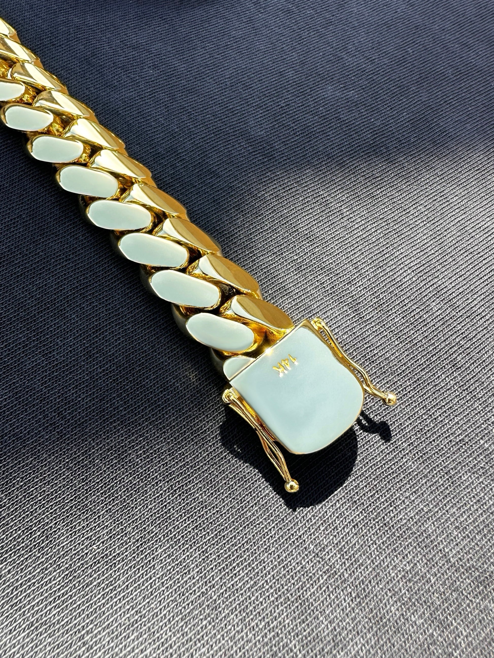 14K gold-plated Miami Cuban link bracelet with box clasp, polished finish, and secure side locks, displayed on dark fabric under natural sunlight.