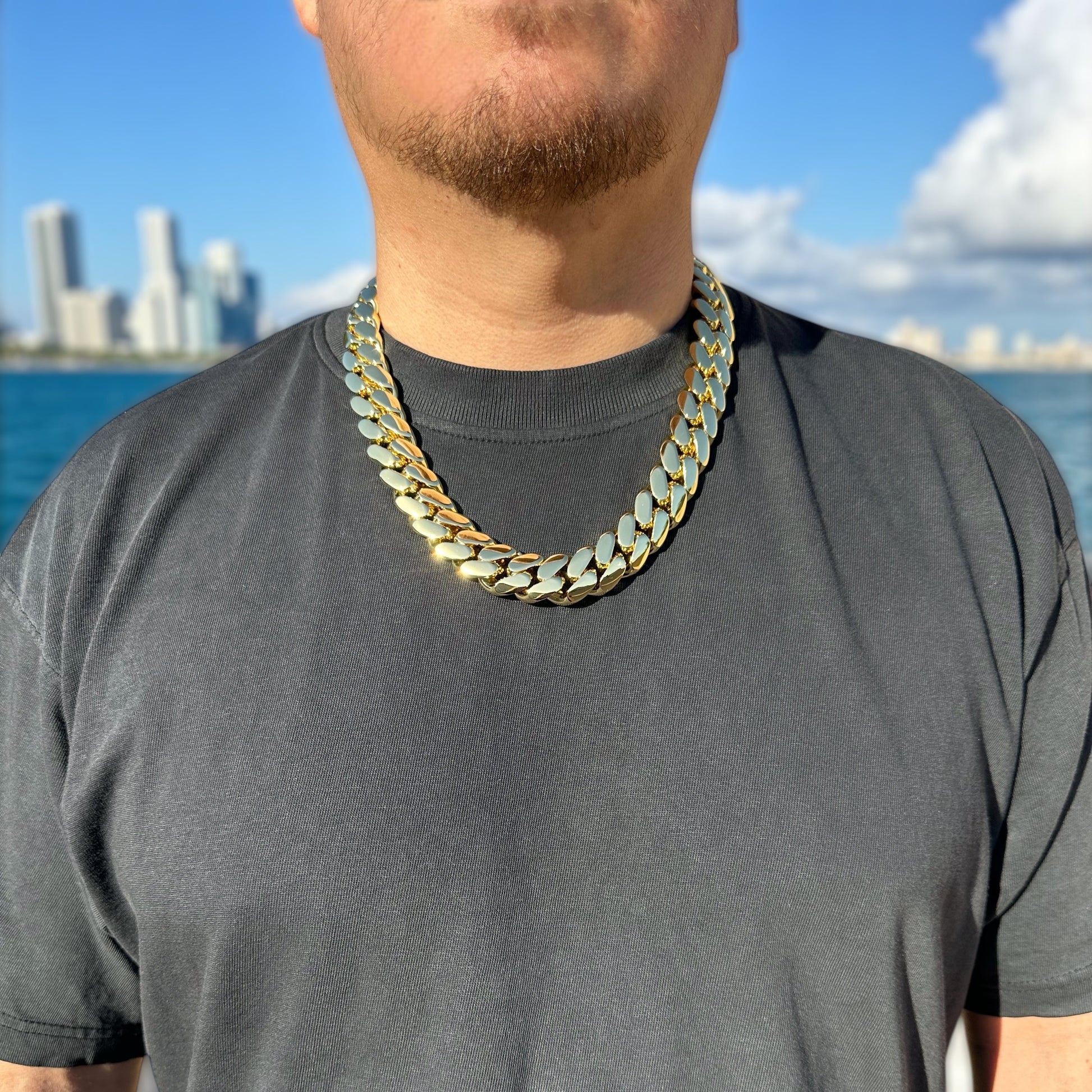 Person wearing a bold 22mm gold chain necklace with a cityscape and water in the background