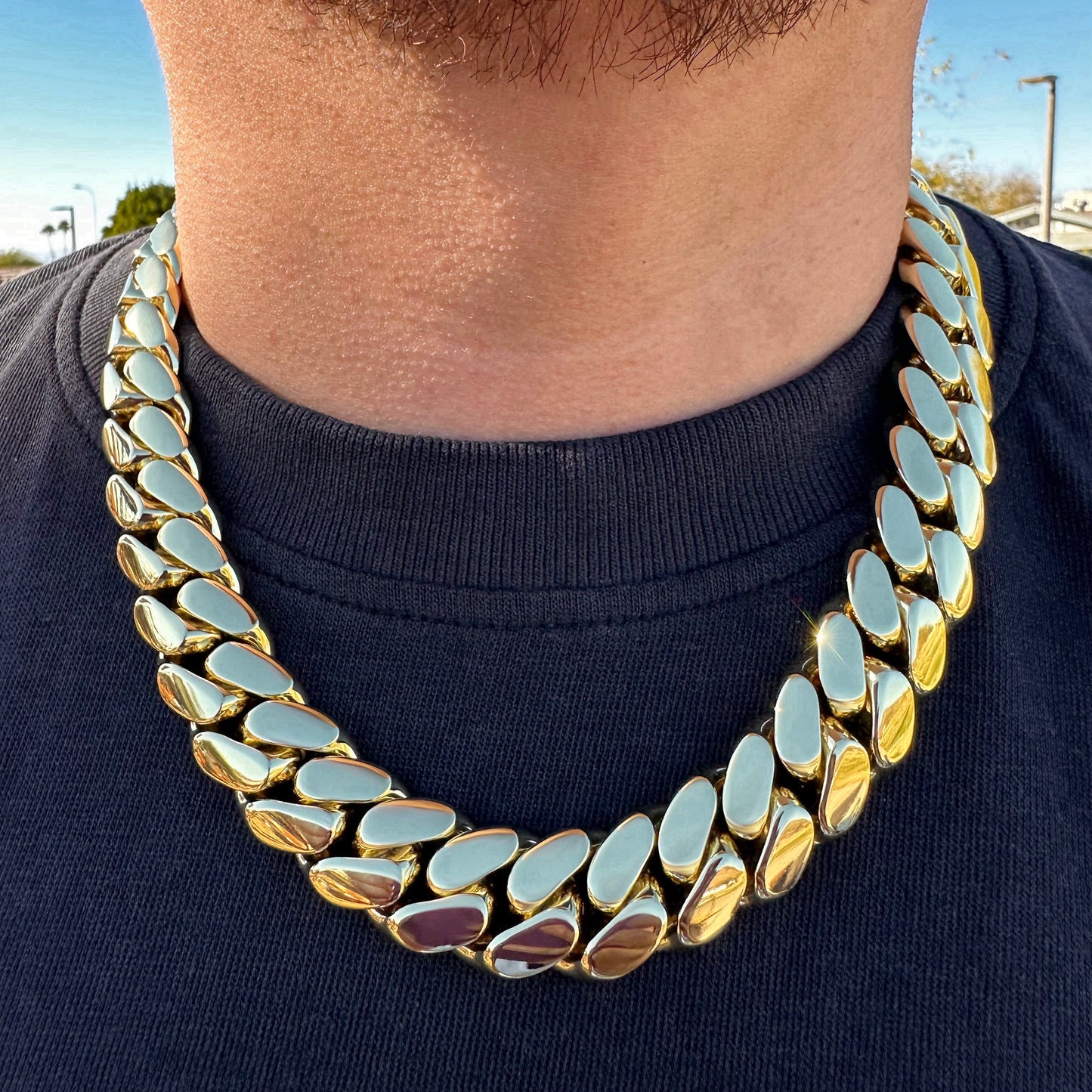 Close-up of a thick 14k gold-plated Miami Cuban link chain on a man’s neck, showcasing the tight links and high-polish mirror finish, worn over a black shirt in natural daylight.
