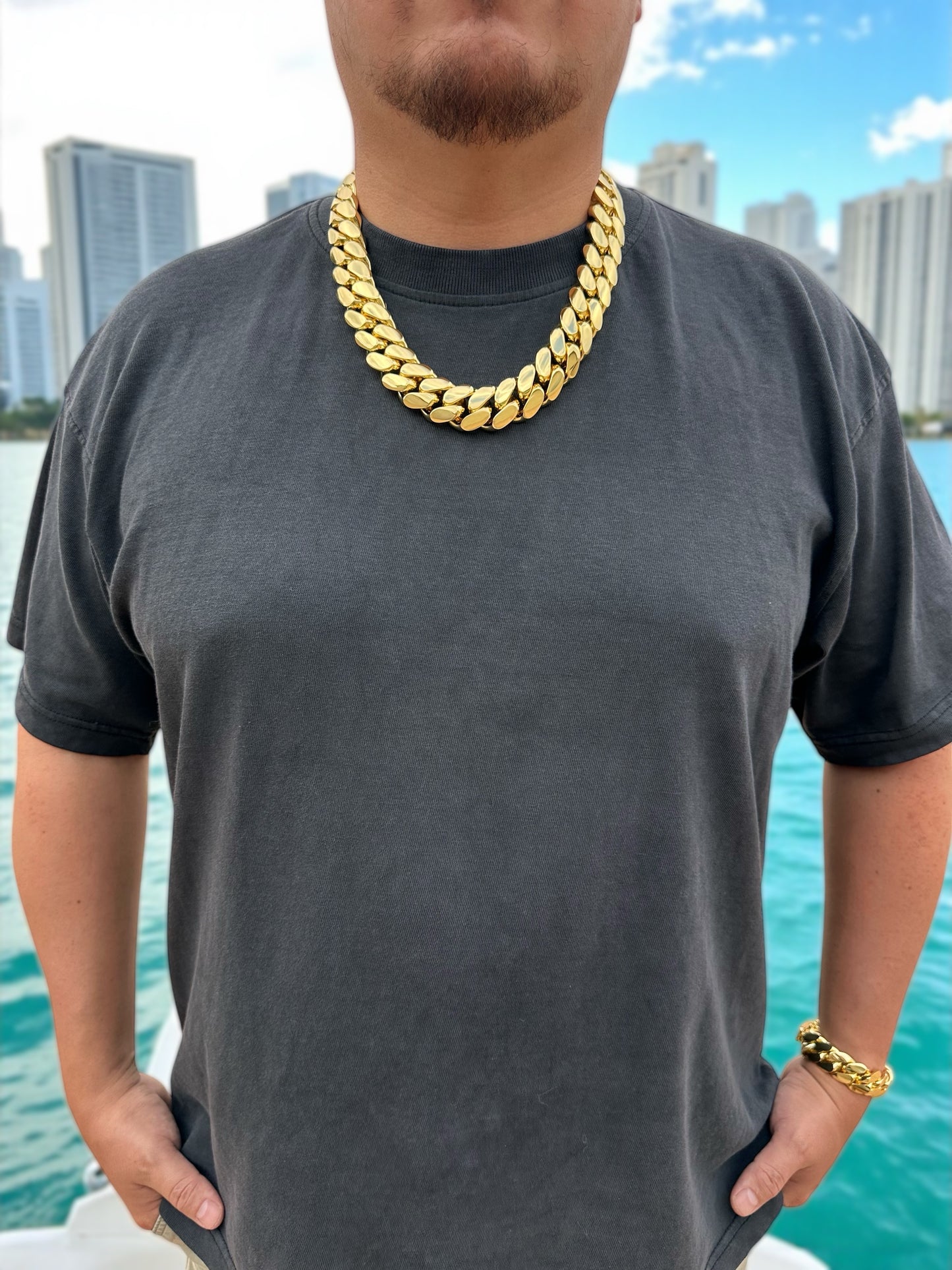 Stylish man wearing a heavy 24mm 14K gold-plated Miami Cuban link chain necklace and matching bracelet, posing on a yacht with the ocean and Miami skyline in the background. Statement luxury streetwear jewelry for men, perfect for hip hop fashion and urban style.