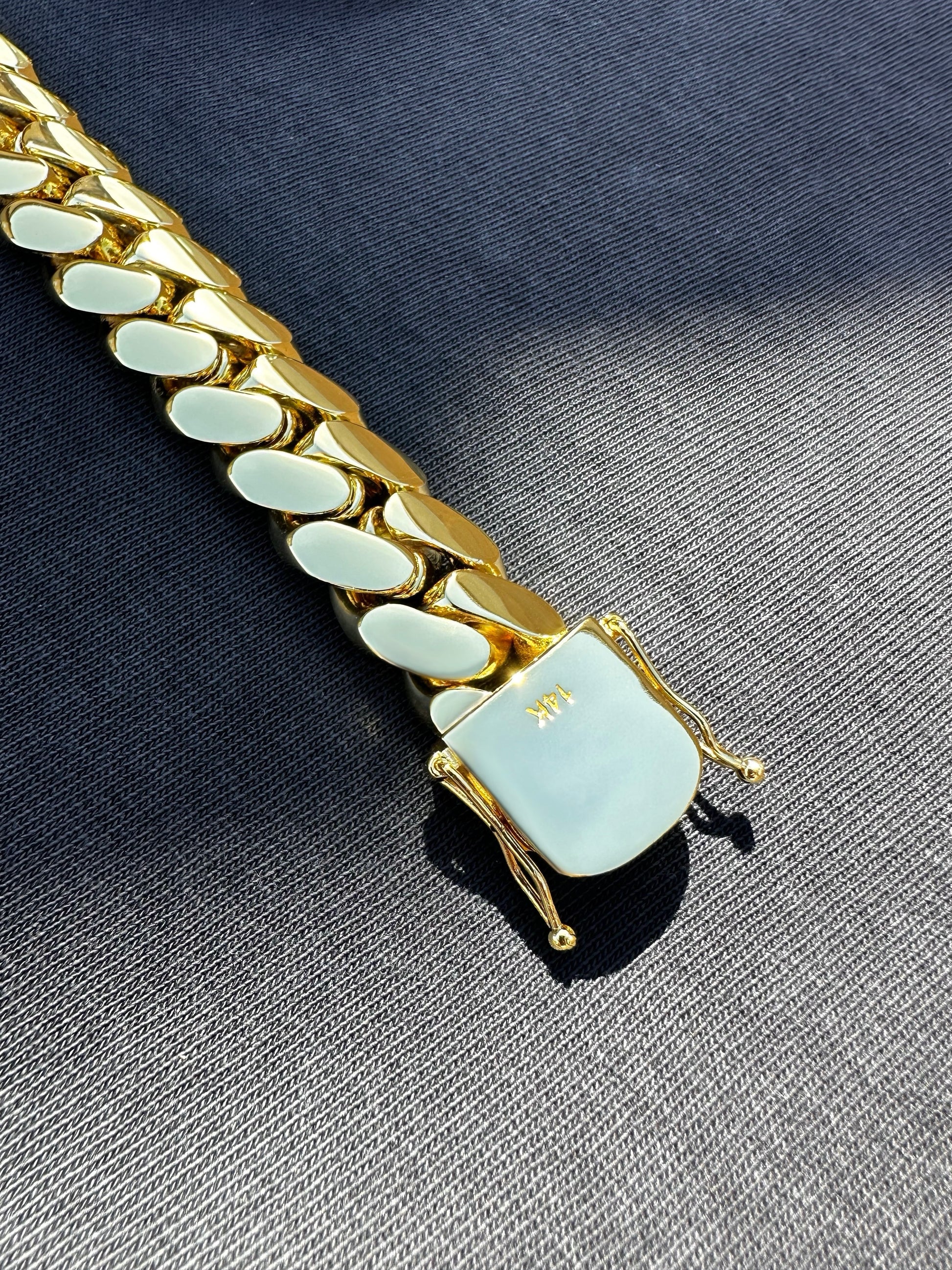 14K gold-plated Miami Cuban link bracelet with box clasp, polished finish, and secure side locks, displayed on dark fabric under natural sunlight.