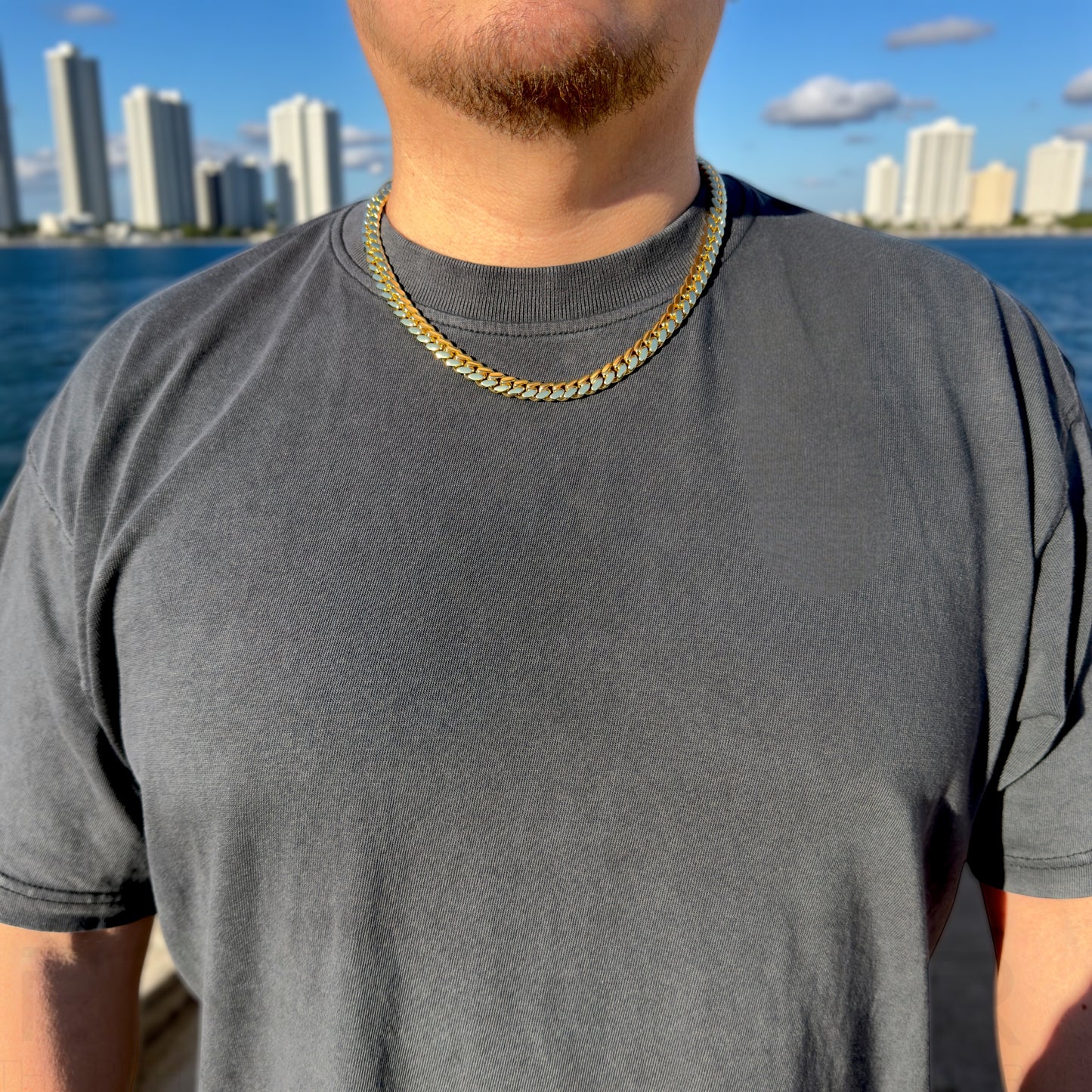 Man wearing a pigment dyed black t-shirt with a 10mm gold cuban chain necklace, standing by water with city skyline in the background.